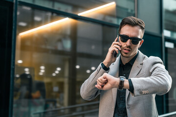 Serious handsome businessman talking on the cell phone while checking the time in modern city street. Busy male person arranging a business meeting.