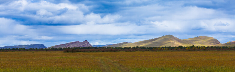 Panoramic landscape with Khakassia steppe at early autumn day in Siberia, Russia. Mountain range Sunduki on the horizon under blue sky. Overgrown dirt road in a field