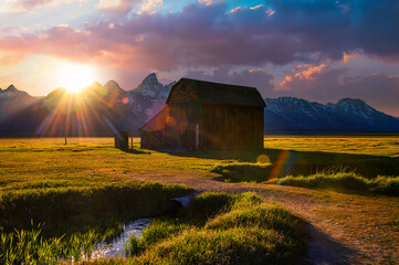 Summer sunset over the historic Thomas Murphy Barn at Mormon Row in Grand Teton National Park, with snowcapped Teton Mountain Range in the background and a water stream in the foreground.