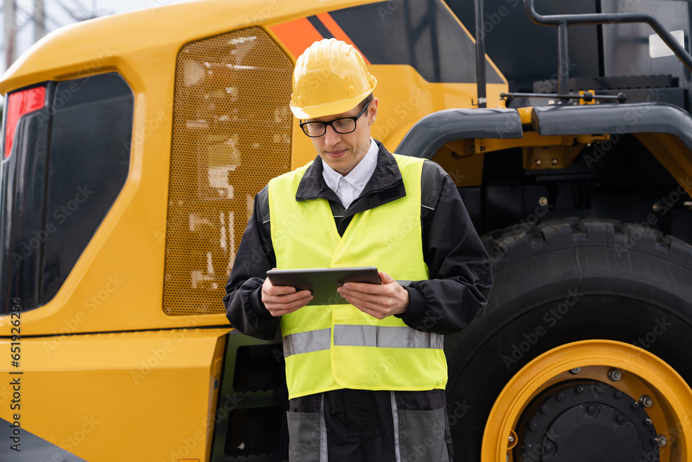 Poster Worker in a helmet with a digital tablet on the background of construction machine