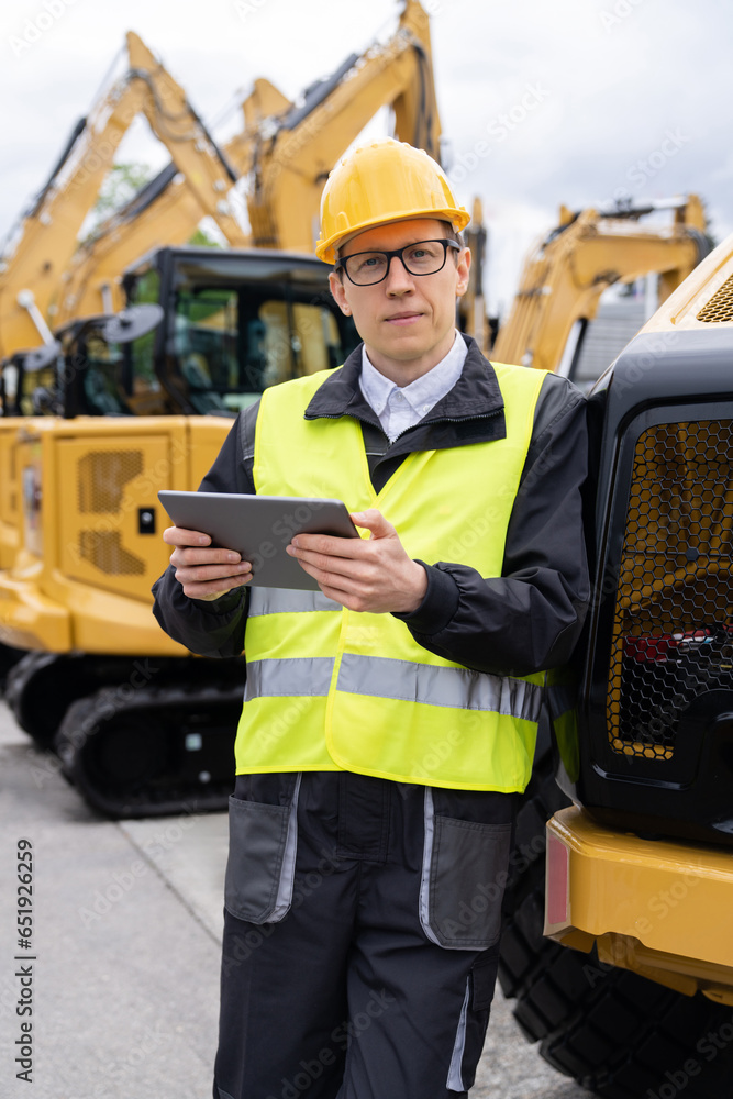 Aufkleber engineer in a helmet with a digital tablet stands next to construction excavators.