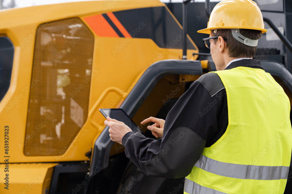 Poster Worker in a helmet with a digital tablet on the background of construction machine