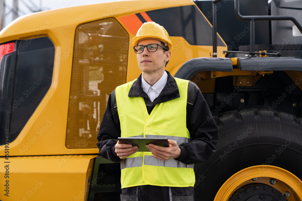 Poster Worker in a helmet with a digital tablet on the background of construction machine