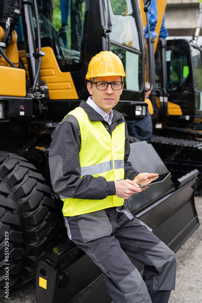 Poster engineer in a helmet with a digital tablet stands next to construction excavators.