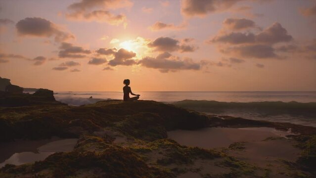 Silhouette of a slim woman on the beach at a beautiful sunset, Practice of yoga breathing techniques. Beautiful purple sunset on bali rocky beach. Female person in lotus position. Sport exercises