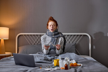 Fototapeta na wymiar young red-haired sick girl holding smart phone sneezing, bless you. closeup portrait, laptop , pills, medicine antiviral antibiotics antipyretic painkillers are on bed