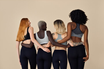 Young diverse fit women wearing sportswear standing on beige. Multicultural girls four models group in tops leggings hugging together on background. Yoga fitness and diversity. Back rear view.