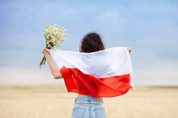 Woman with bouquet of daisies holding flag of Poland in wheat field. Back view. Polish Flag Day. Independence Day. Travel and learn polish language concept.