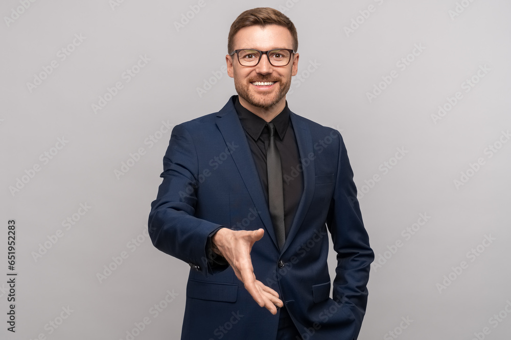 Wall mural happy businessman holds out hand for handshake looking at camera on gray background. man in suit smi