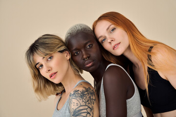 Three cool confident pretty gen z girls looking at camera posing for beauty portrait, multiethnic stylish young women, multicultural hipster models inclusive faces isolated on beige background. - obrazy, fototapety, plakaty