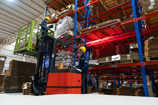 Male warehouse worker using forklift electric or electric pallet stacker lift lifting female colleague to check cardboard boxes or corrugated paper sheet goods on shelves in store warehouse.