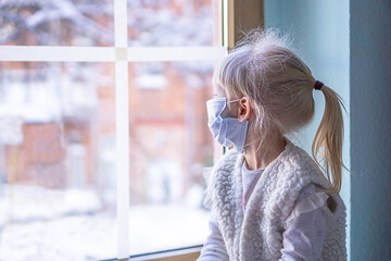 Little girl in mask sitting in front of window and looking out to snow tree branches. 