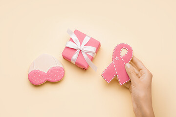 Female hand holding pink cookie in shape of ribbon, heart with bra and gift box on beige background. Breast cancer awareness concept