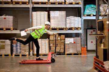 joyous caucasian male staff dressed in working clothes and helmet in warehouse having fun standing on pallet truck jack, driving among shelves, enjoy free time, leisure at work place
