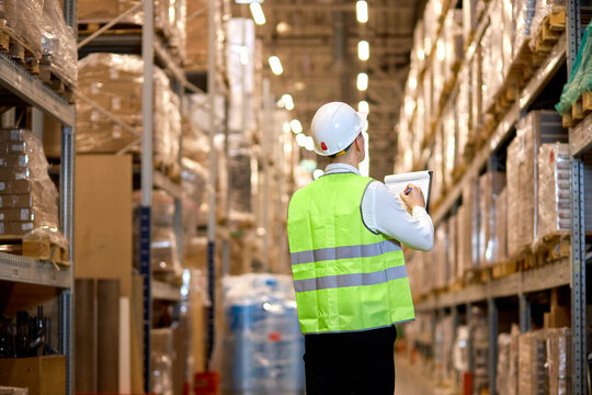 rear view on young male in helmet with clipboard checking goods at warehouse, counting and controlling distribution. unrecognizable hardworking caucasian worker in green working uniform