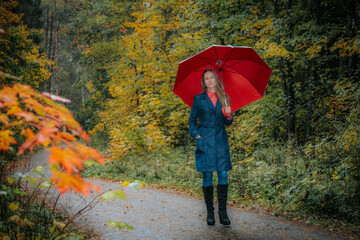 Joyful Woman Finds Shelter Under Vibrant Umbrella in Autumn Forest. Happy young woman in autumn forest, surrounded by colorful foliage and trees. Soft selective focus 