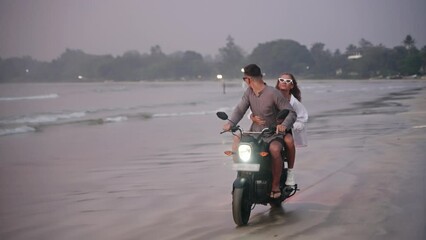 Travelling couple rides motorbike on sea beach on tropical island at sunset. Man, woman travel by motorcycle on coastline along ocean. Lady hugs boyfriend on adventure. Motivational background.