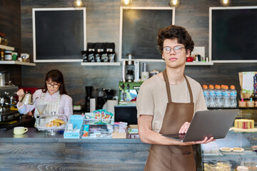 Young guy in an apron with laptop in his hands inside coffee shop cafeteria
