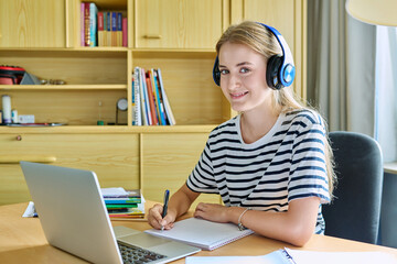 Girl teen student in headphones sitting at desk using computer writing in study notebook