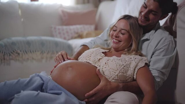 Young cheerful Caucasian romantic pregnant couple lying comfortable on living room floor at home. Happy man loving caressing belly of blonde woman in advanced stage. Joyful people expecting a baby.