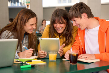 Three smiling Caucasian students snacking drinks and doing homework using tablet in university...
