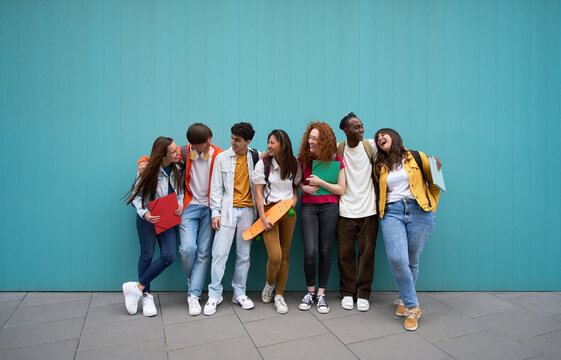 Large group of cheerful young people leaning against blue wall. Multicultural college students enjoying campus recreation. Happy friends hugging and laughing. Generation z and international relations.
