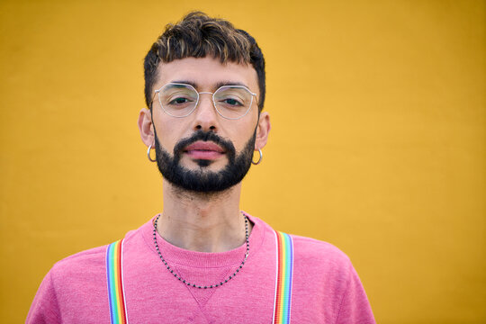 Close up portrait attractive serious young Caucasian man rainbow LGBT suspenders. Gay millennial people posing looking camera for isolated photo yellow background. Earnest person confident studio.