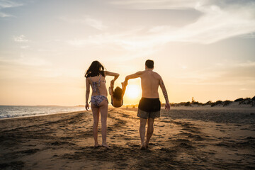 young couple sunbathing their son on the beach, playing in the sunset light - obrazy, fototapety, plakaty