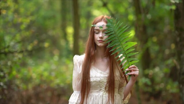 Beautiful red haired girl with tiara in medieval dress on glowing sun. Fairy tale story about princess.Amazing model in forest holding fern.Warm art work