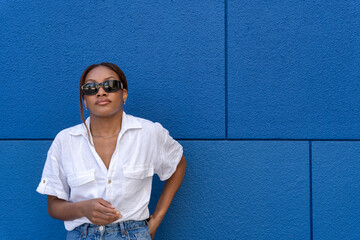 Pretty young woman of black ethnicity with sunglasses looking at the camera and white shirt, on a blue background.