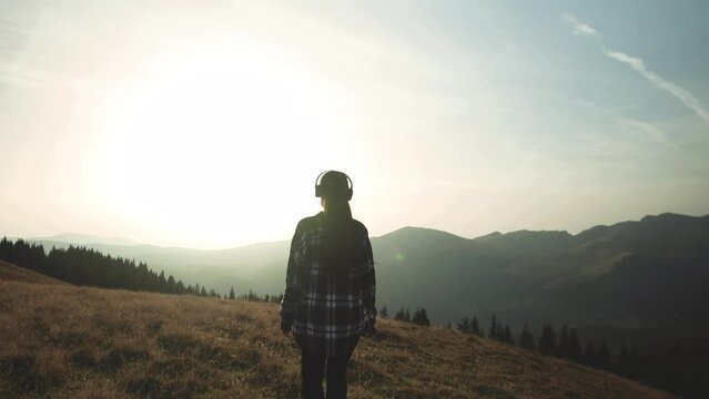 Young Woman Walking On A Hill With Headsets Listening To Music And Relaxing. Female Hiker Walking Towards Sunset In Mountain Environment In Early Autumn. Tracking Slow Motion Shot From The Back 