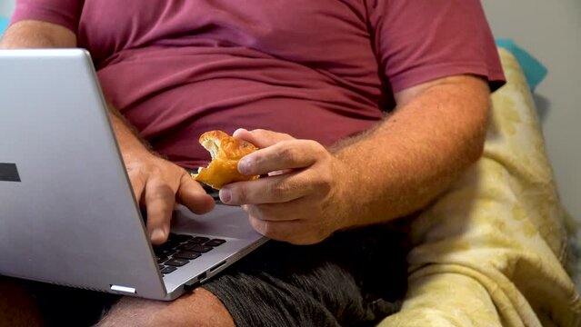 Sedentary overweight man, sitting eating pastries next to a phone and a laptop.