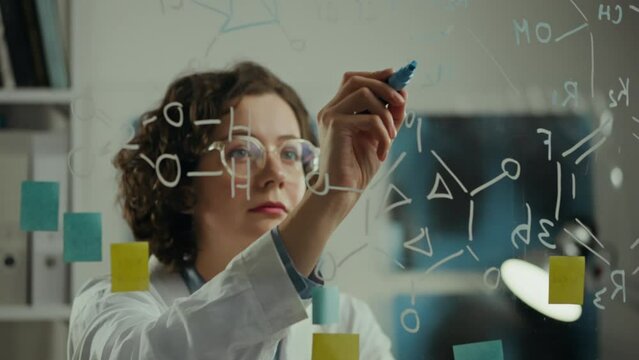 Specialist. Female chemist tensely writes formula using a glass wall in scientific institute. Camera in motion Skilled worker Working with data on transparent wall Creating study strategy, test tube