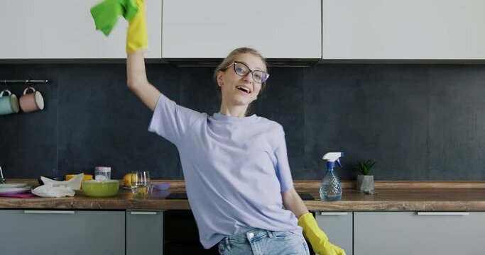 A cheerful young girl with glasses dances and wipes a plate while cleaning the kitchen. Good mood while cleaning.