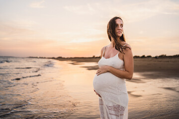 portrait of beautiful pregnant woman in white dress, on the sea shore with sunset light