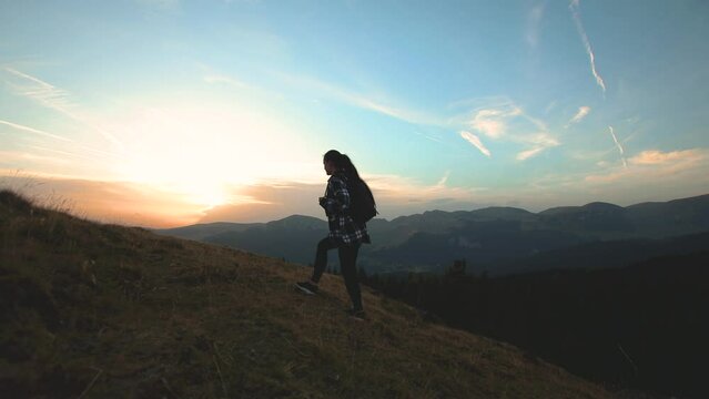 Young woman hiking with backpack. Girl climbing a hill with sunset and mountains landscape in the background. Female hiker walking and relaxing in the mountains environment