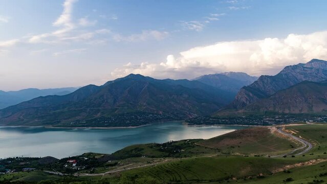 Mountains and lake video timelapse for background. Light percy clouds quickly fly in blue sky over mountains and lake or Charvak reservoir. Day turns into night. Village lights on shore of lake