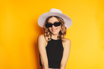 Close-up portrait of blonde woman casual portrait in positive view, big smile, beautiful model posing in studio over yellow background. Caucasian portrait woman in white hat and black top, eyeglasses.