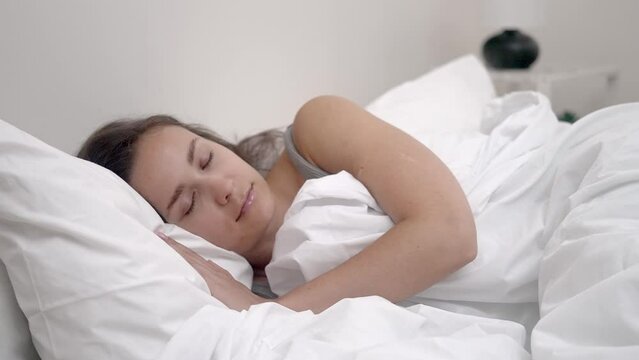 Tranquil Slumber: A young woman enjoys peaceful sleep in a bright bedroom, embraced by a soft white blanket and a comfortable orthopedic mattress. 