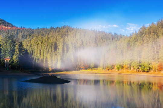 autumn landscape with lake among the coniferous forest. sunny morning with blue sky and mist above the water surface