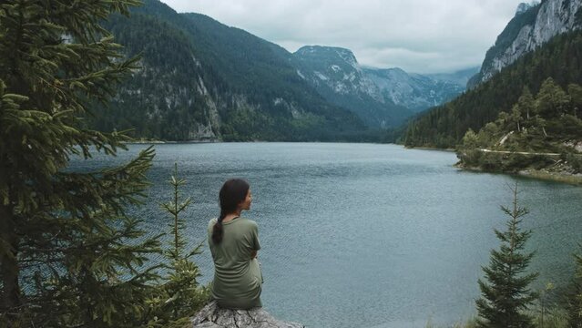 Young Woman Standing back in Forest and looking at mountain lake landscape. She Enjoys Gentle Breeze, Breathes Deeply and Smiles Subtly. Slow Motion Cinematic Shot. Tranquility and peace concept