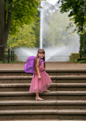 A little smiling girl in a pink beautiful dress with long hair with a backpack goes to school, photo on the steps in the park. Against the background of a fountain
