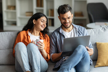 Young hindu couple resting on sofa with laptop, woman drinking coffee, spouses resting in living room, browsing internet together, choosing movie for evening - Powered by Adobe