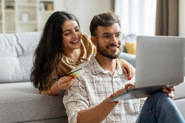 Indian couple with laptop computer shopping online from home, paying for order in internet, purchasing and buying things, sitting in living room