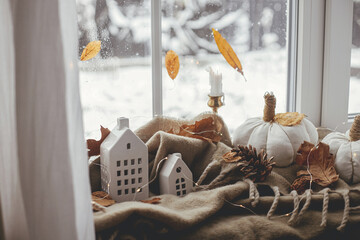 Cozy Autumn. Stylish pumpkin pillows, fall leaves, candle, lights and cute buildings decoration on brown scarf on windowsill. Autumn hygge, fall home decor. Happy Thanksgiving