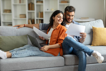 Happy indian couple checking documents, spouses reading insurance agreement or property certificate, husband and wife sitting on couch in living room, copy space - Powered by Adobe