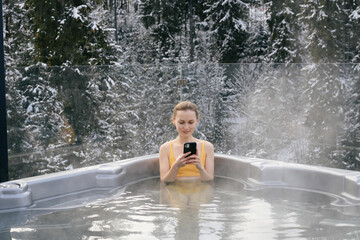 A young woman is looking at a mobile phone and relaxing in the hot water of a jacuzzi outdoors in winter. The concept of outdoor recreation and recovery during vacation - obrazy, fototapety, plakaty
