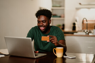 Fototapeta na wymiar Portrait of a happy african american man sitting at home and using a credit card and a laptop for online shopping.