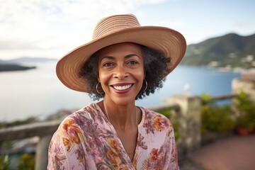 Fototapeta premium A middle-aged woman wearing a half-hat smiles and looks at the camera on the shore of the ocean and mountains. Vacation, travel.