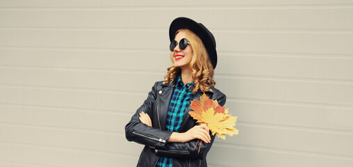 Autumn portrait of stylish happy smiling young woman with yellow maple leaves wearing round hat and rock style black leather jacket posing on gray background